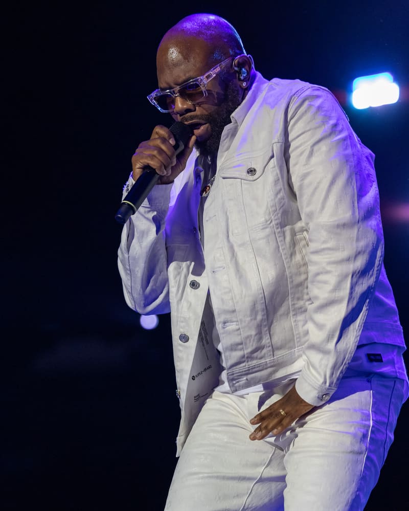 Performer singing into microphone on stage wearing white jacket, sunglasses and purple stage lighting.