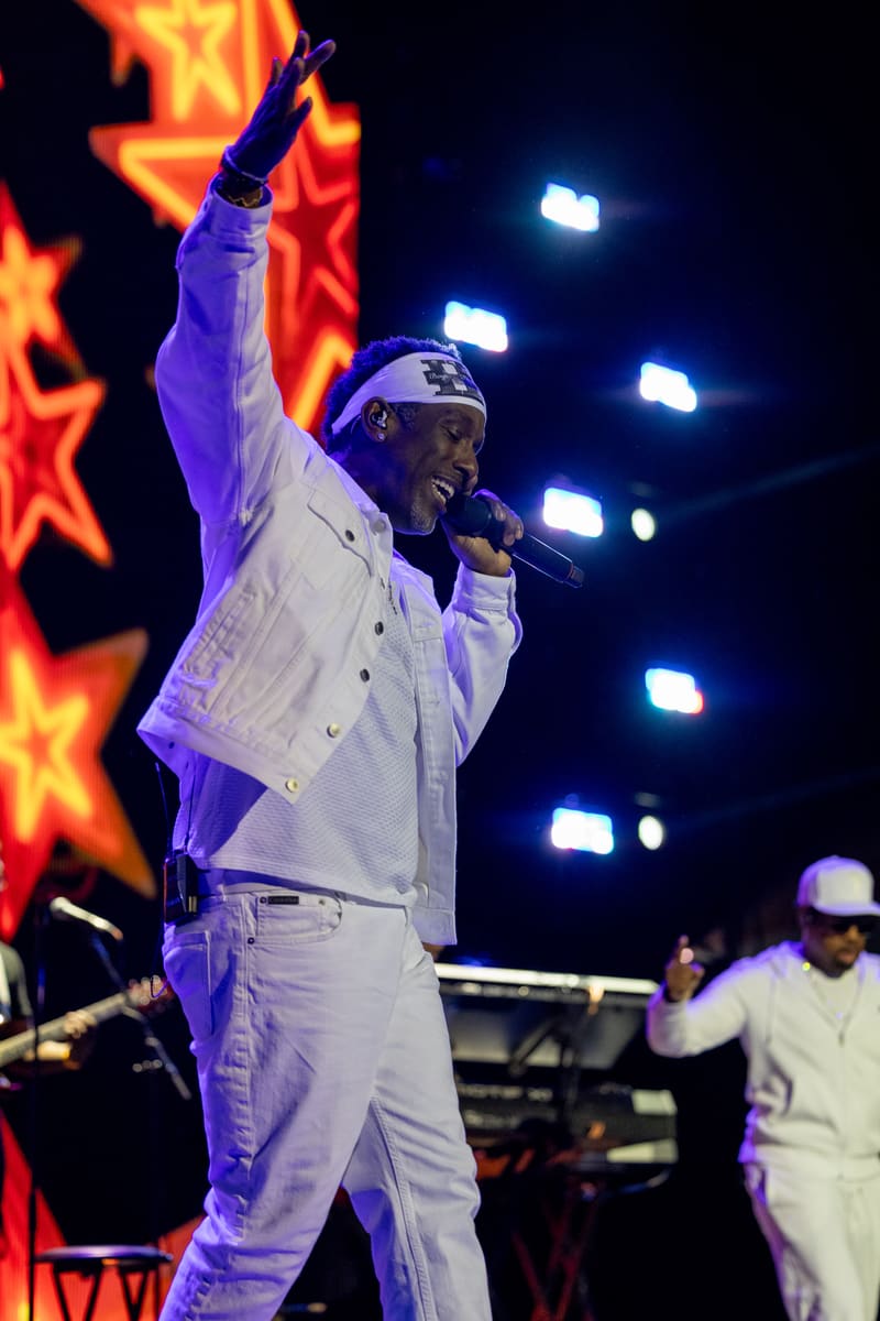 Performer in white suit and cap singing into microphone with fist raised, illuminated by stage lights and fiery backdrop graphics.