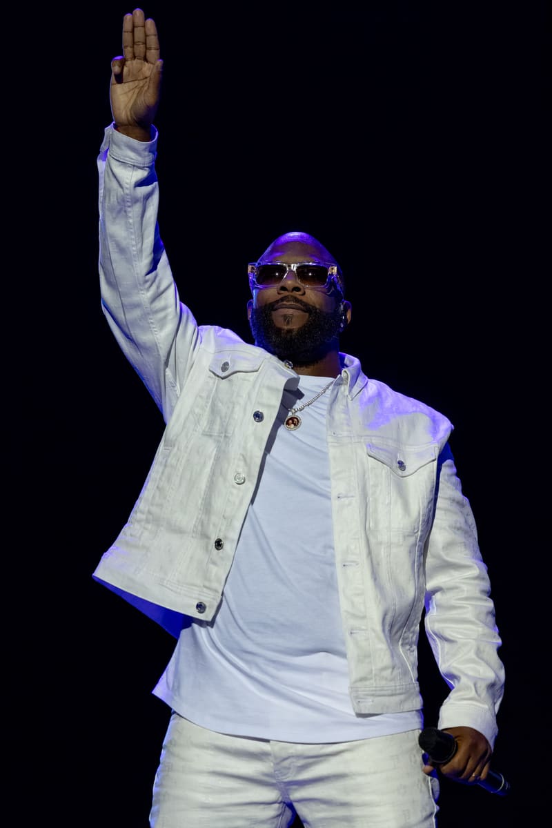 Performer in white jacket and sunglasses raises fist to crowd under purple stage lighting.