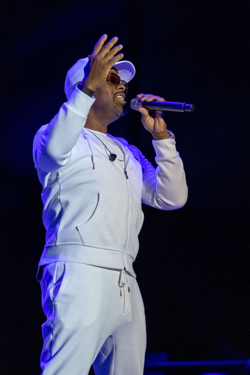 Male singer in white tracksuit and sunglasses performing at microphone under purple stage lighting.
