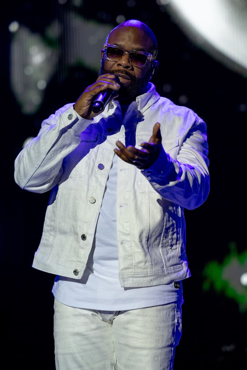 Male performer singing into microphone wearing white jacket and sunglasses, lit by purple stage lighting.