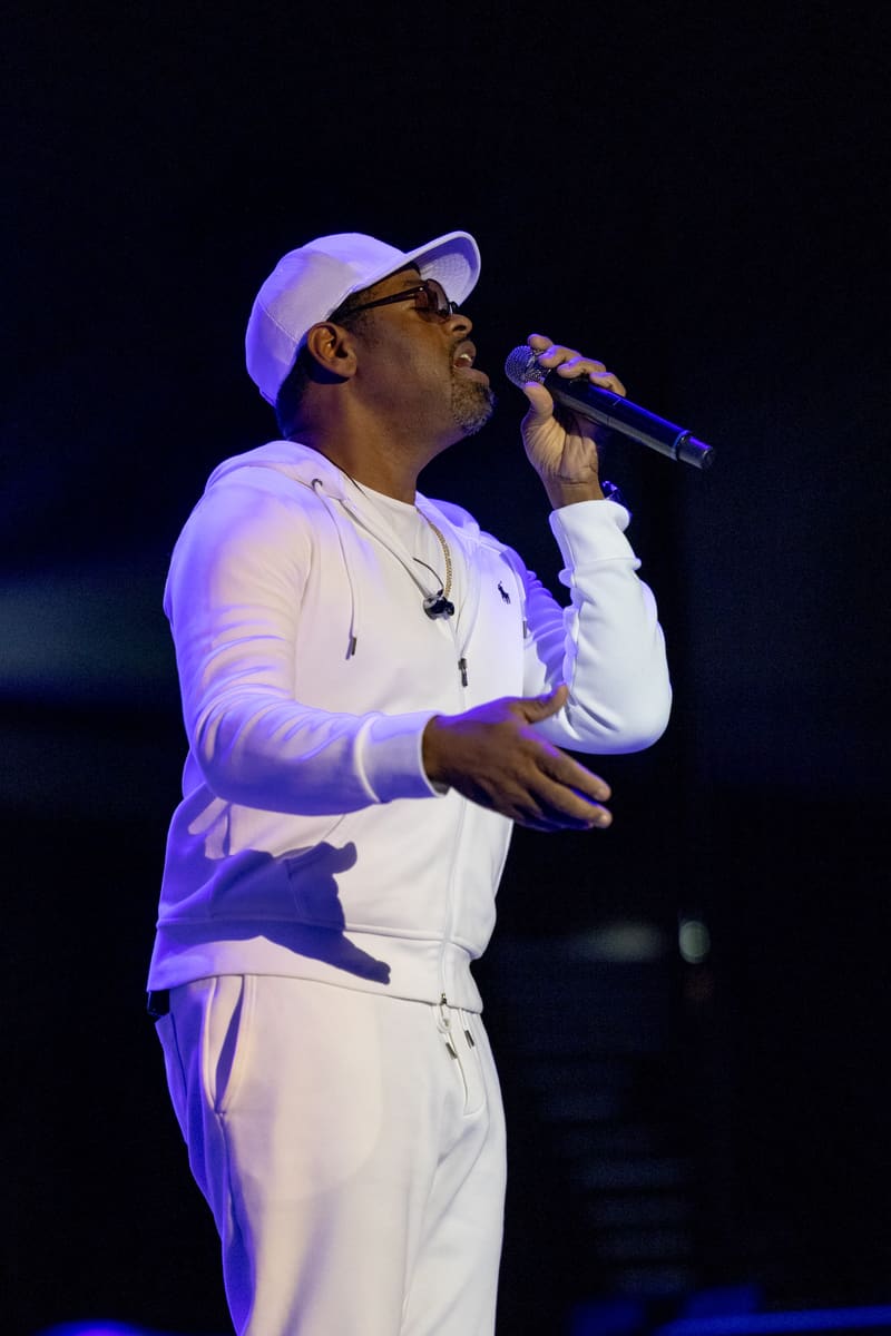 Singer in white tracksuit and cap performing at microphone under purple stage lighting.