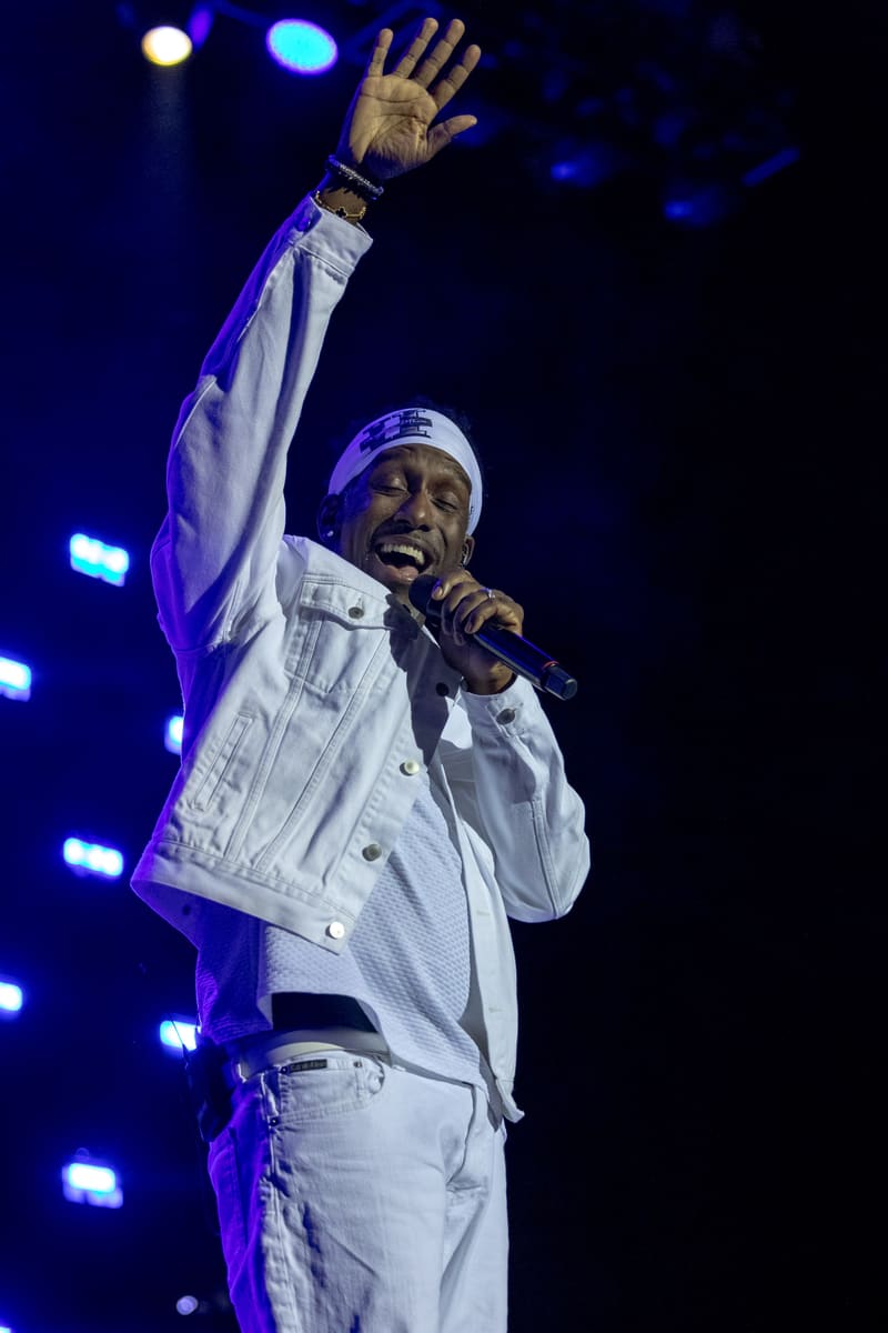 Performer in white suit and cap raises fist while singing into microphone under blue stage lights.