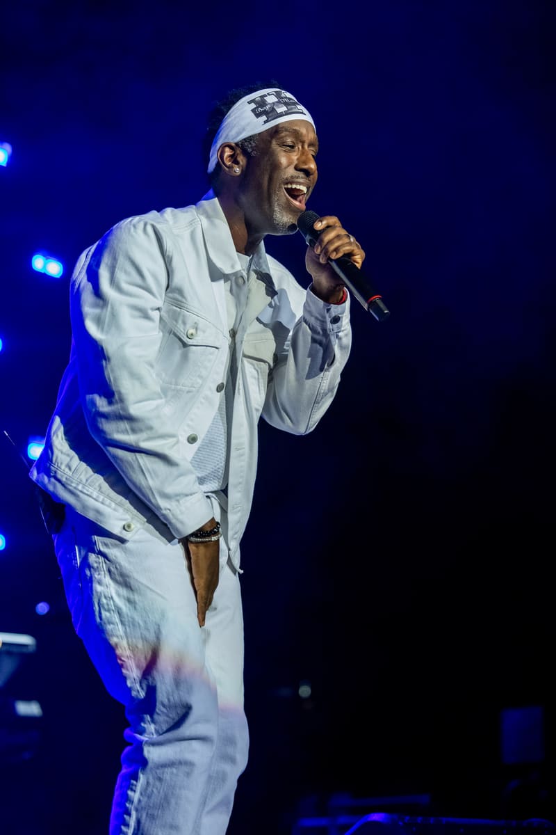 Singer in white outfit and headwrap performing with microphone under blue stage lighting.