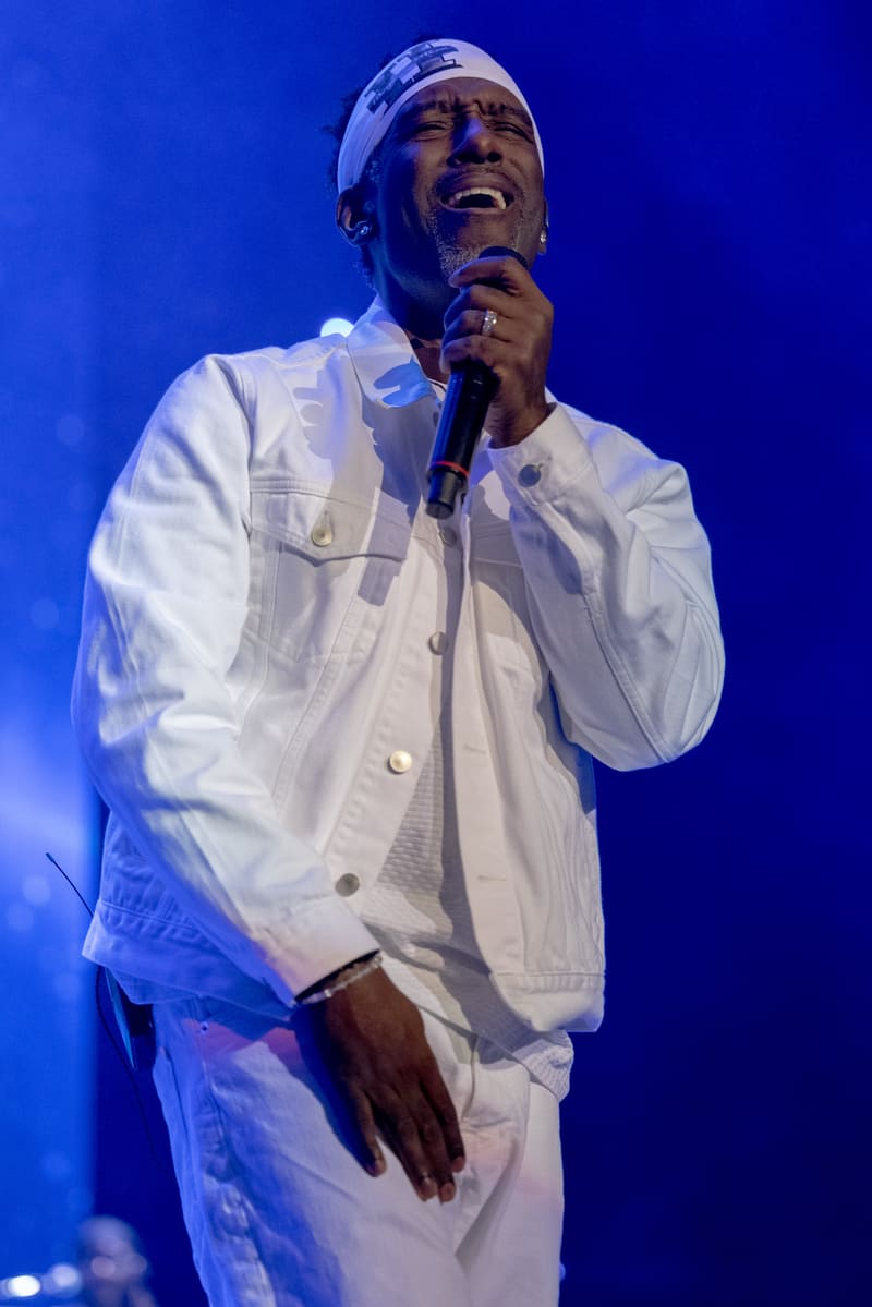 Singer in white suit and headwrap performing with microphone under blue stage lighting.