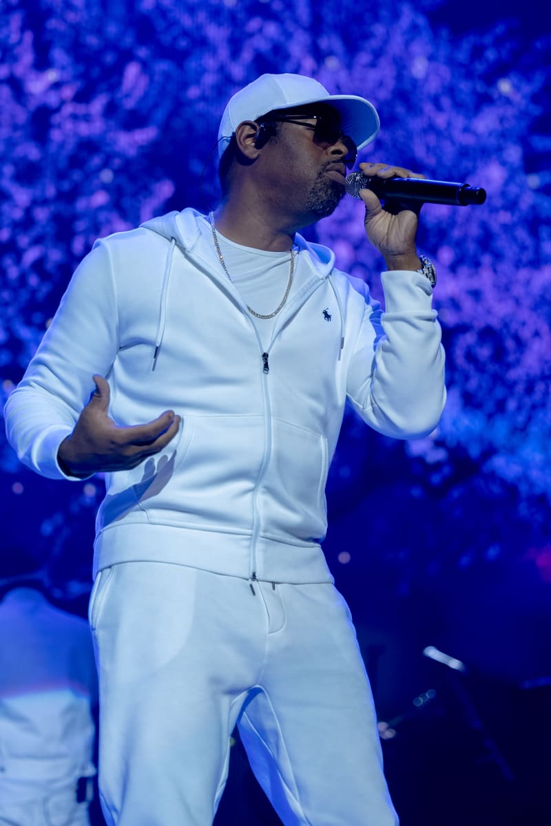Performer in white tracksuit and baseball cap singing into microphone against blue-lit stage backdrop.