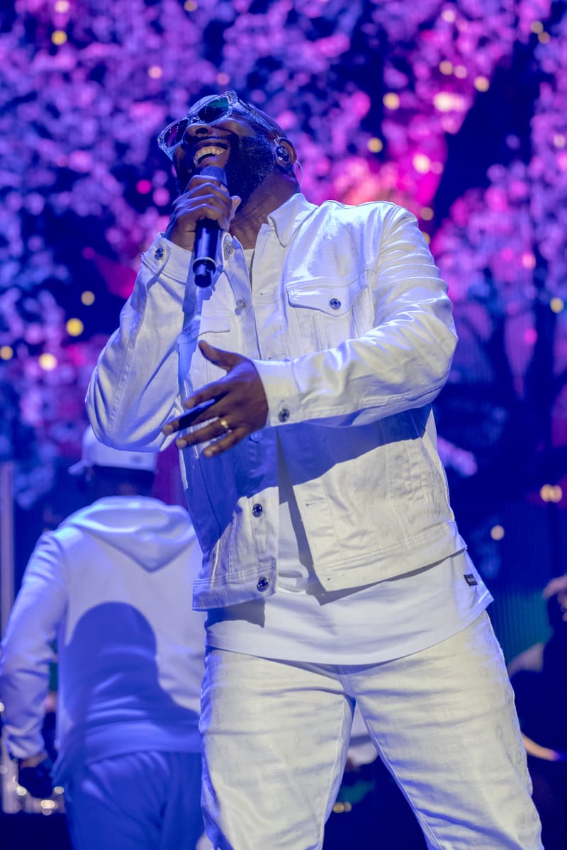 Singer in white suit performing with microphone against vibrant purple-lit stage backdrop.