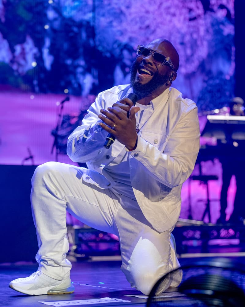 Performer in white suit and sunglasses kneeling on stage with microphone, bathed in purple stage lighting.