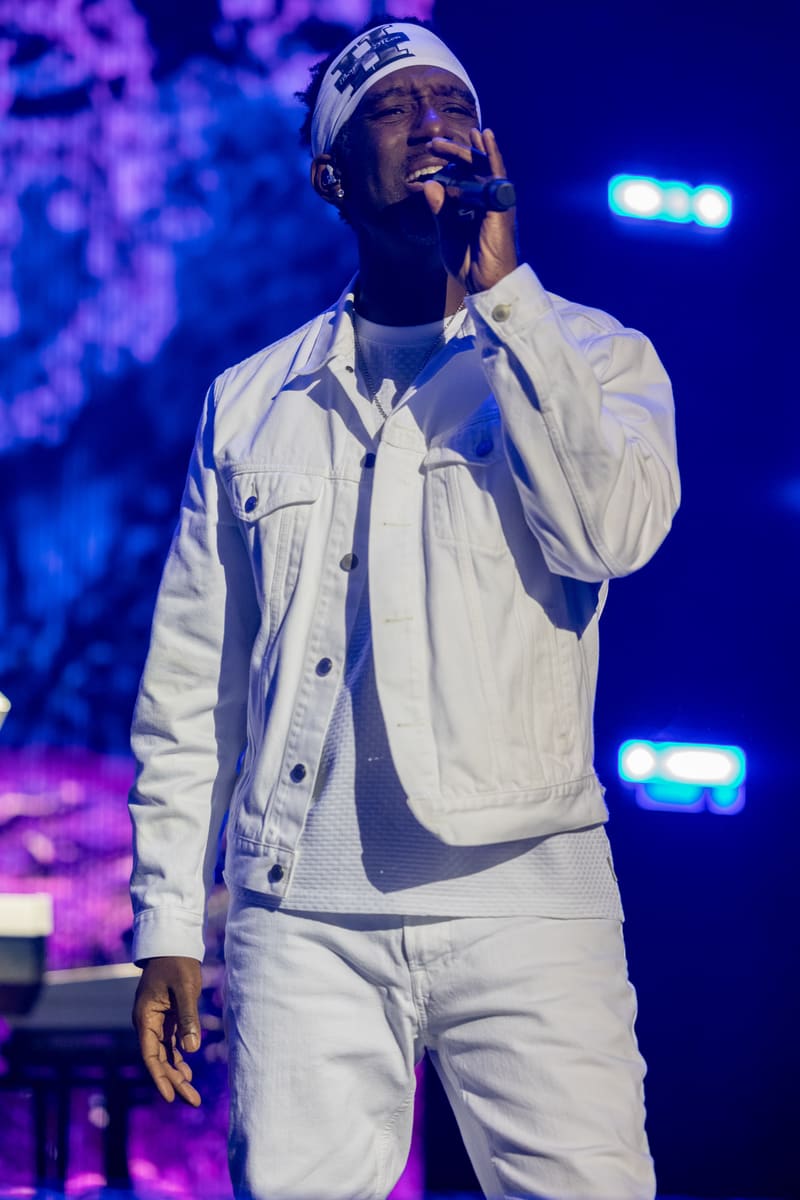 Male performer singing into microphone wearing white shirt and headband, lit by blue and purple stage lighting.
