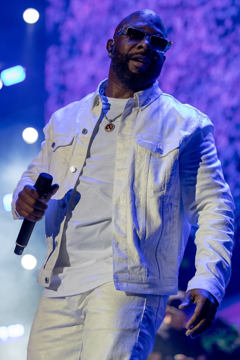 Performer in white denim jacket and sunglasses holding microphone on stage bathed in blue stage lighting.