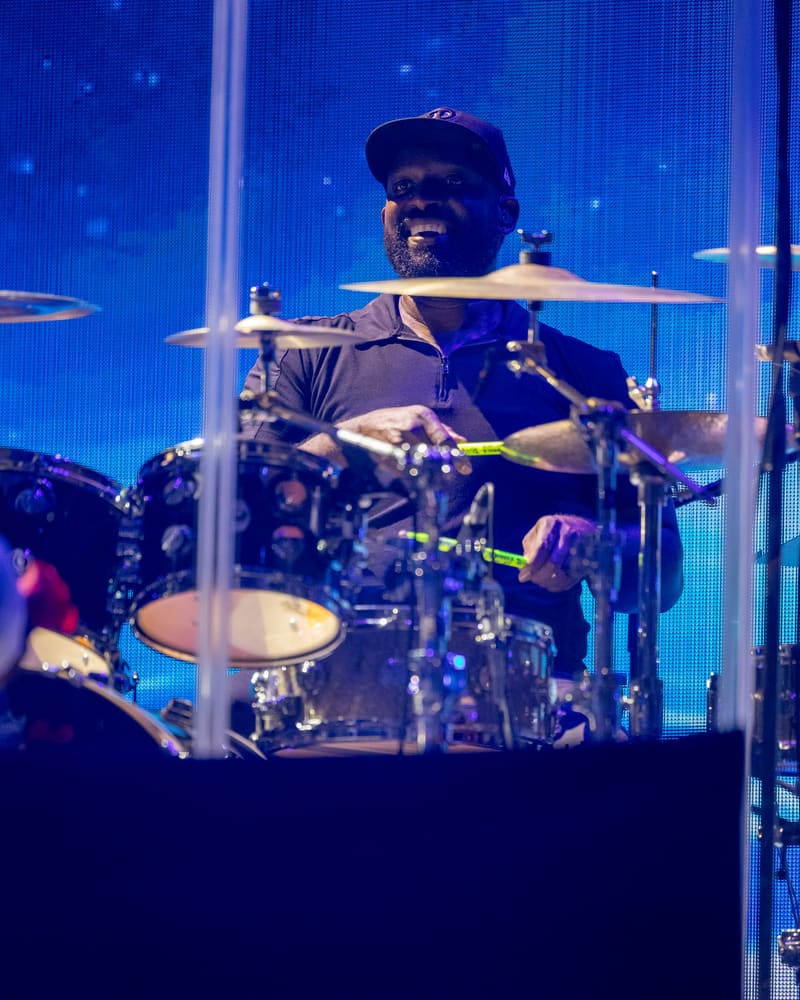 Drummer performing at drum kit against blue-lit stage backdrop during Fridayz Live concert.