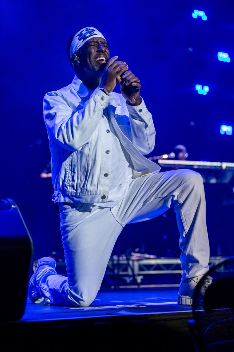 Male performer kneeling on stage in white suit and headwrap, holding microphone under blue stage lighting.