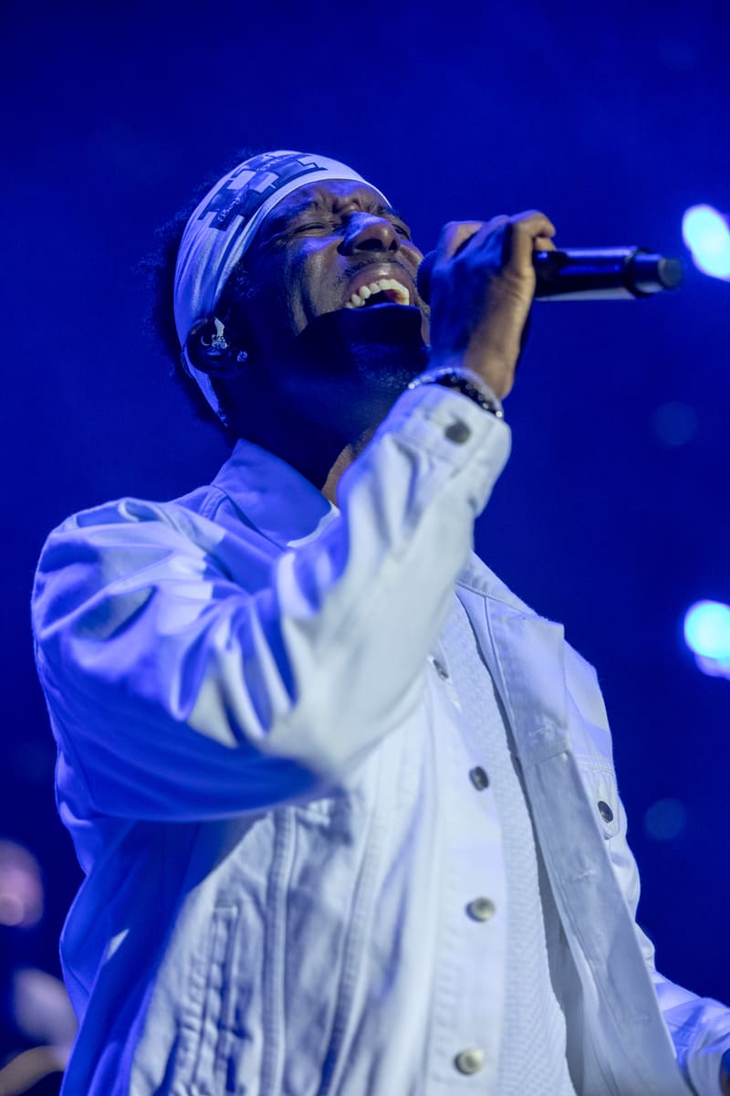 Male performer singing into microphone on stage wearing white shirt and head wrap, lit by blue stage lighting.