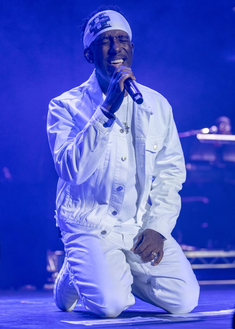 Singer in white jacket and headband kneeling while performing into microphone under blue stage lighting.
