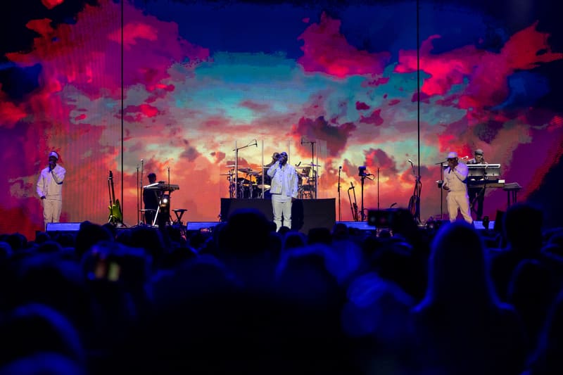 Band members perform on stage beneath a vibrant sunset-coloured backdrop during a live concert, with audience silhouettes in the foreground.