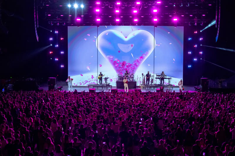 Large crowd facing a concert stage with a glowing heart symbol on the main screen, surrounded by purple lighting and stage equipment.