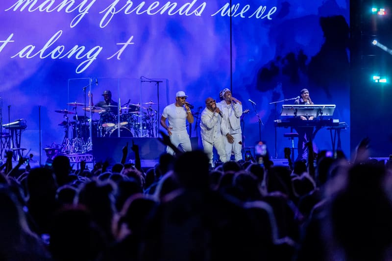 Four performers in white shirts sing on stage under blue lighting as audience members watch from below.