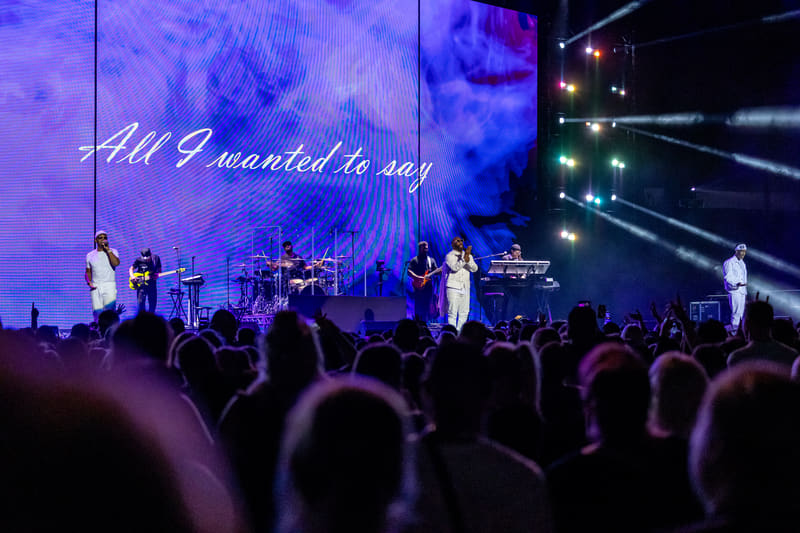 Three performers on stage with band under purple lighting, crowd silhouettes in foreground at Fridayz Live.