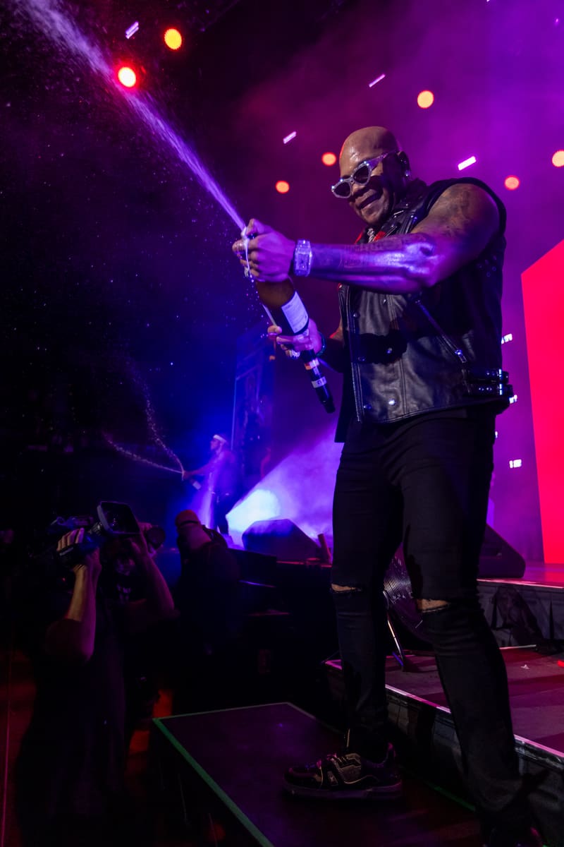 Performer on stage at Fridayz Live lit by purple and blue spotlights, holding microphone with audience visible below.