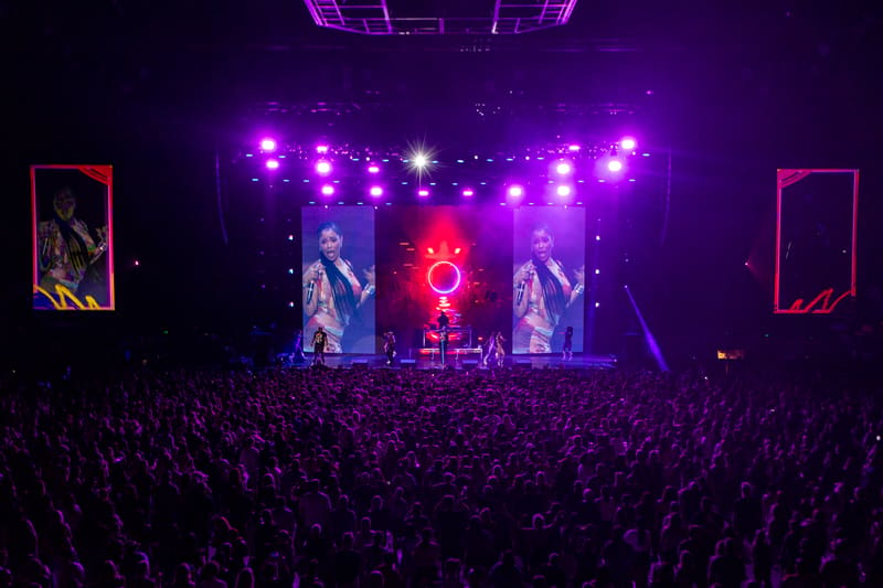 Performer on stage beneath purple and white stage lights with large video screens and crowd in foreground at concert venue.