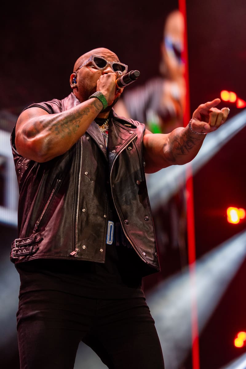 Performer singing into microphone wearing sunglasses and leather vest on stage with red lighting and band members behind.