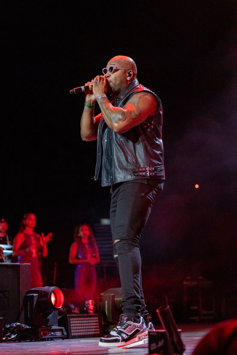 Performer singing into microphone on stage under red stage lighting with band members visible in background.