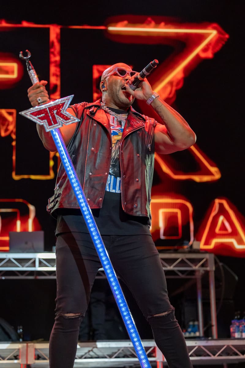 Performer in red leather vest singing into microphone against illuminated neon signage during Fridayz Live concert.