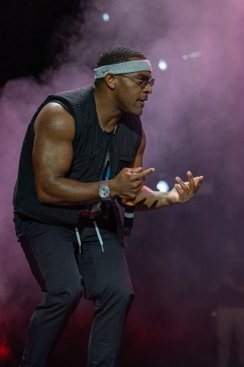 Performer on stage wearing a headband and sunglasses, gesturing expressively amid purple stage lighting and smoke.