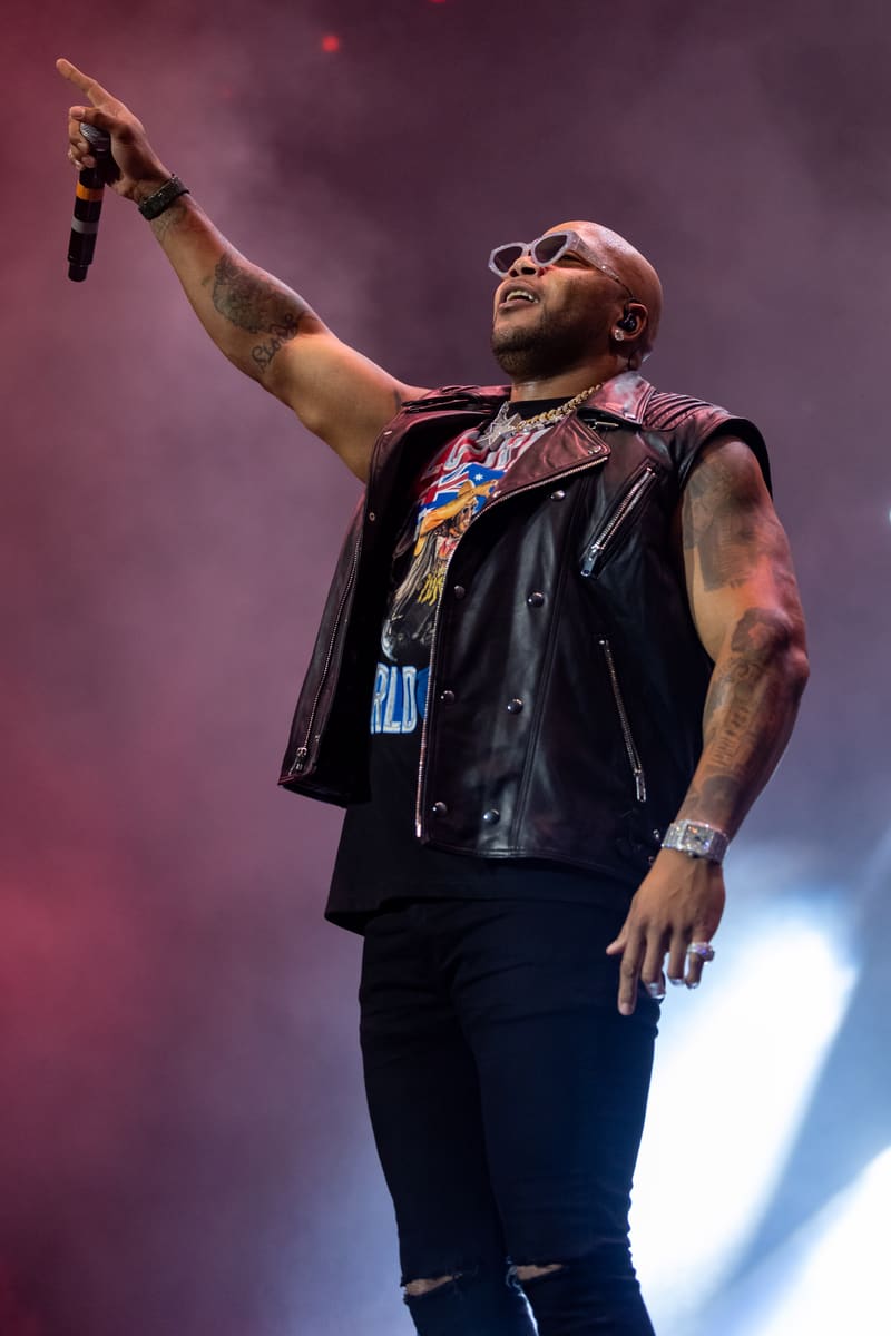 Performer in black leather vest and sunglasses raises microphone arm against purple stage lighting.