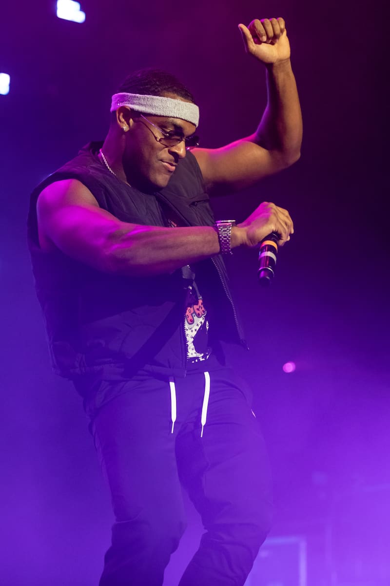 Performer on stage wearing sunglasses and headband, holding microphone with fist raised, bathed in purple stage lighting.
