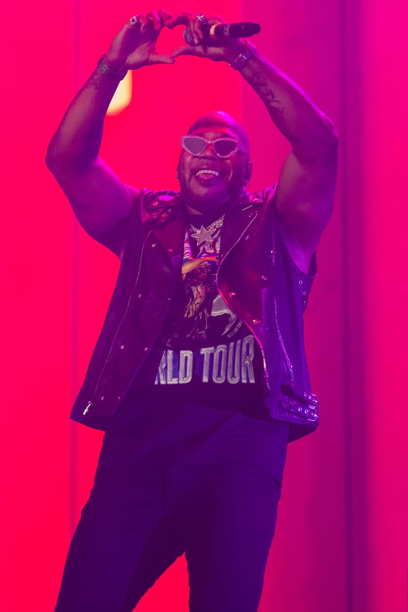 Performer wearing sunglasses and a tour jacket makes a heart shape with their hands under vibrant pink stage lighting.