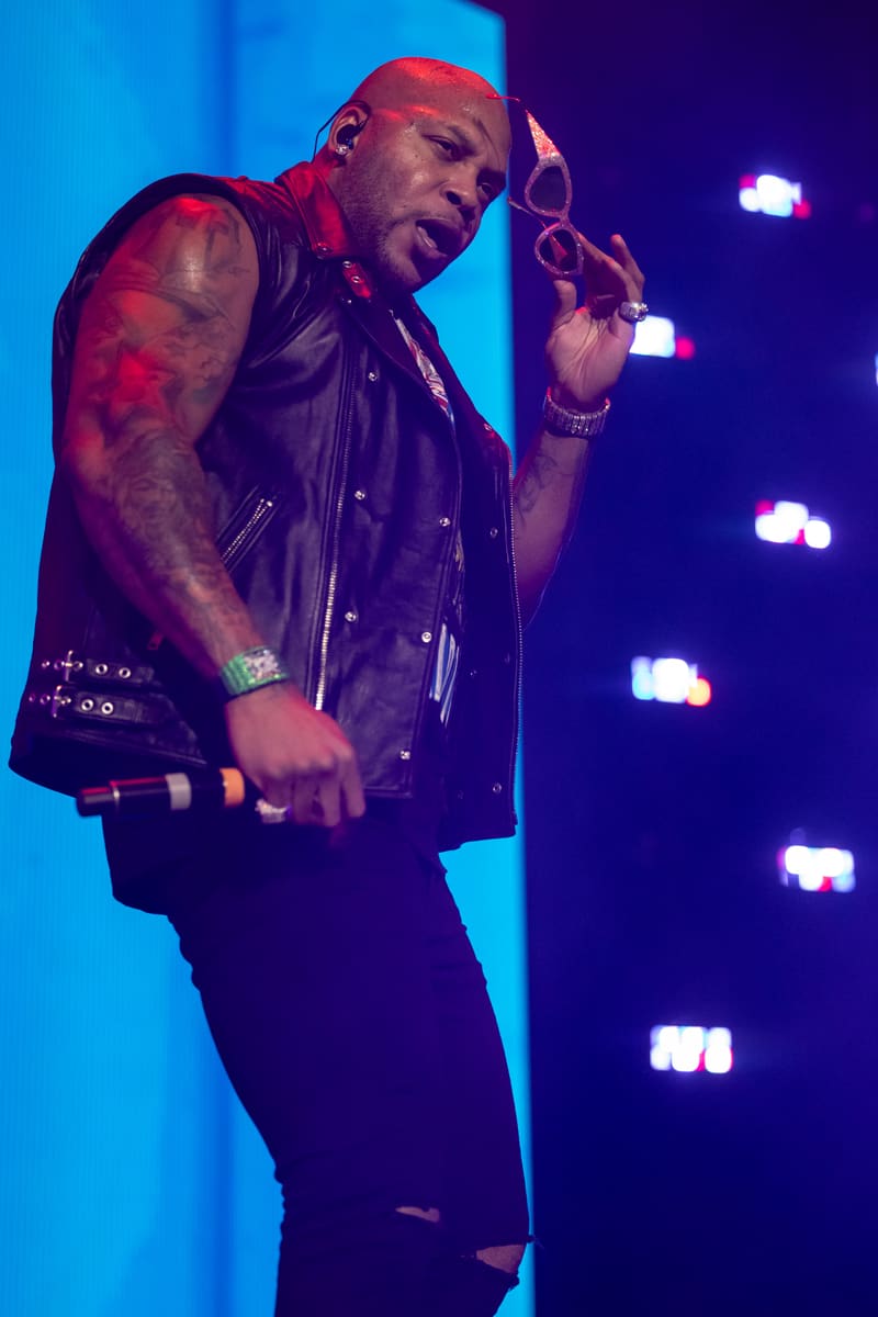 Performer on stage in black leather vest and dark pants, illuminated by blue lighting and stage lights, gesturing expressively.