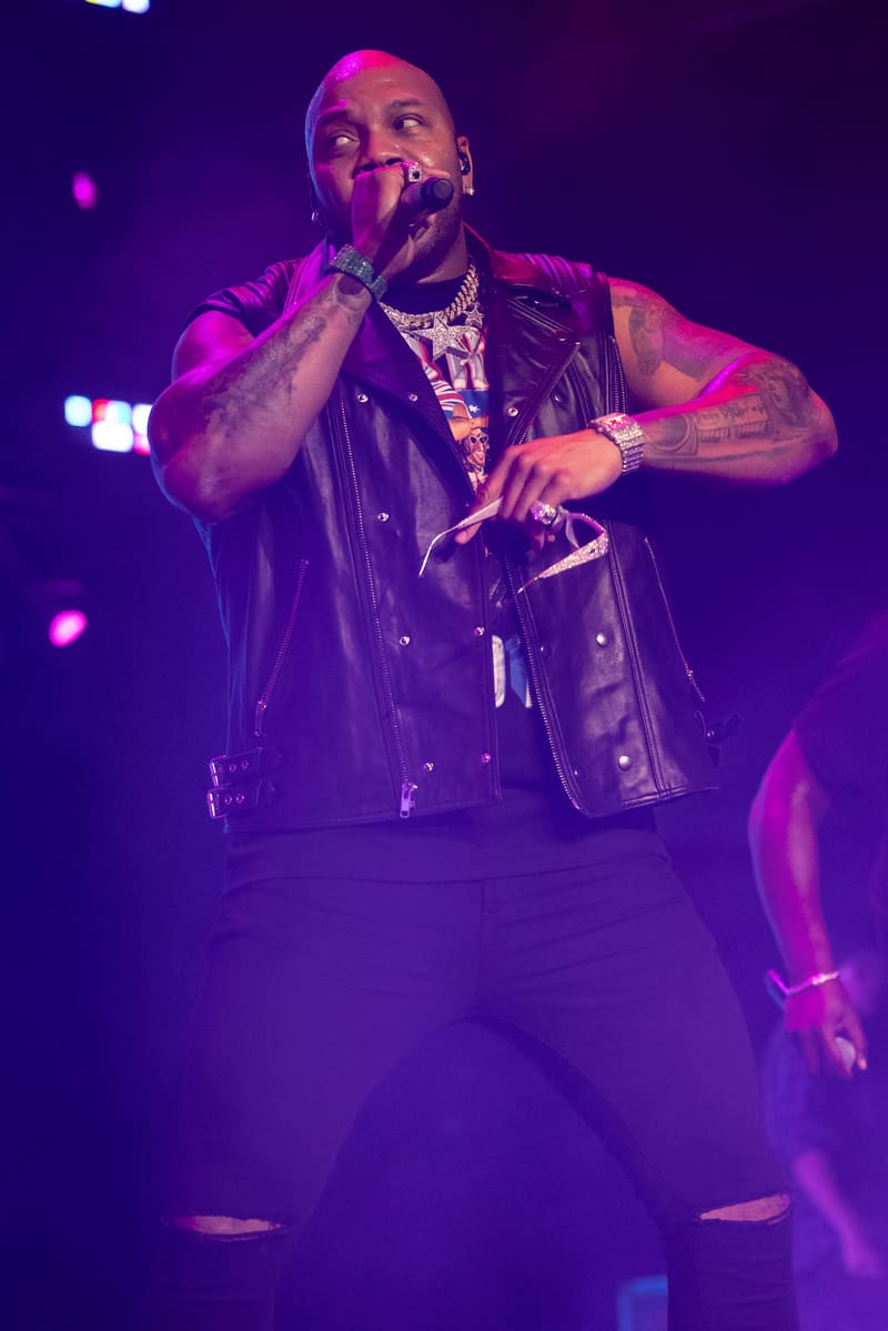 Performer raps into microphone on stage wearing black leather vest, lit by vibrant purple and magenta stage lighting.