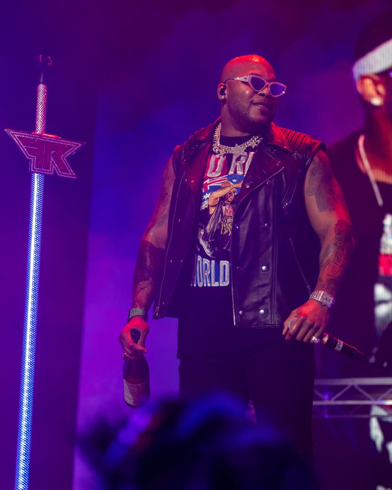Performer on stage wearing sunglasses and a black leather vest, lit by purple stage lighting and a neon star prop.