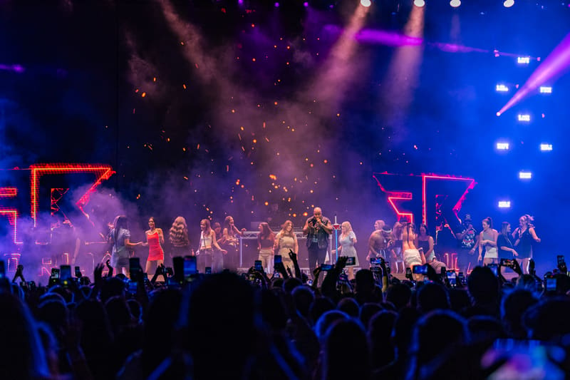 Live band performs on stage beneath purple and blue spotlights, with audience silhouettes in foreground.