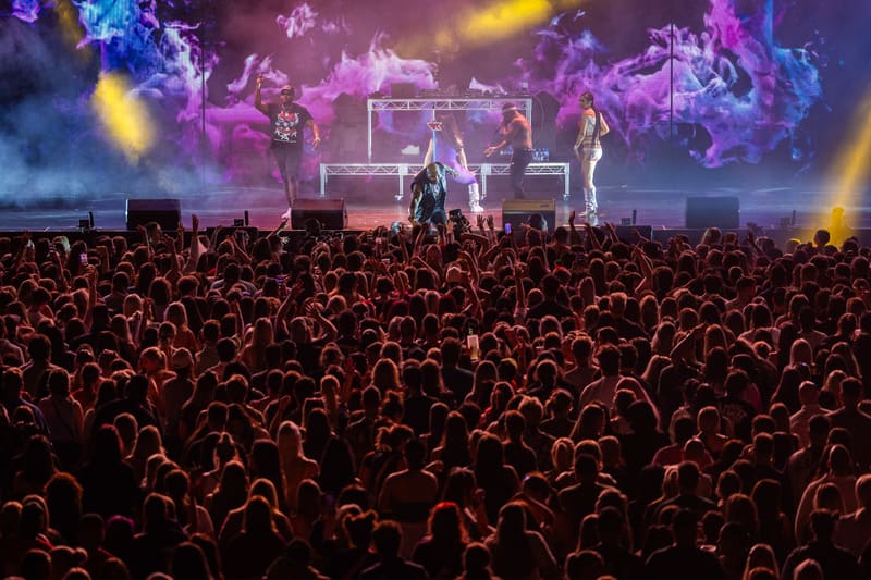 Performers on stage bathed in purple and blue lighting above a large crowd with raised hands during Fridayz Live.