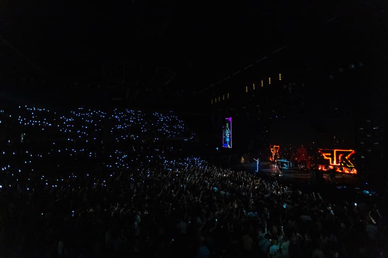 Large crowd gathered under night sky at outdoor music festival with illuminated stage and signage.