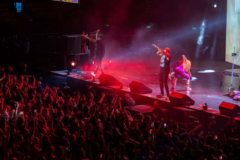 Performers on stage bathed in red and purple lighting with a crowd of audience members in the foreground during a live concert.
