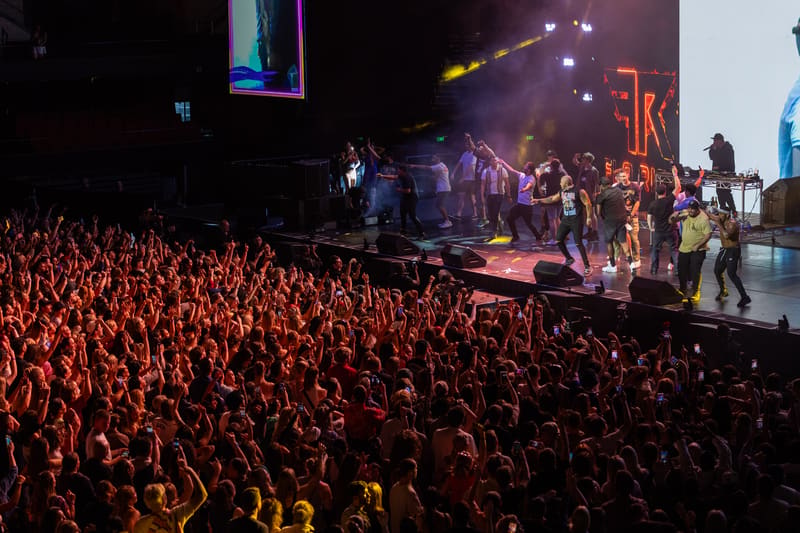 Performer on stage bathed in warm spotlights, surrounded by a dense crowd of thousands with hands raised.