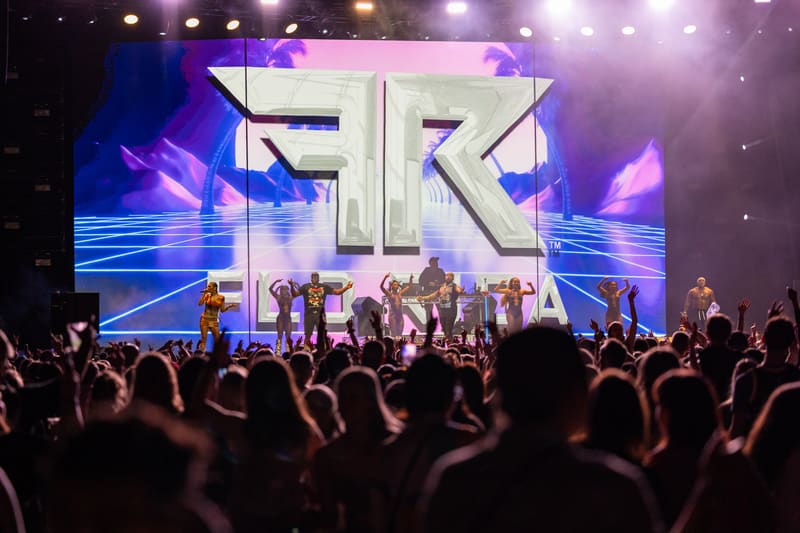 Crowd of music fans with raised phones watch performers on stage beneath large illuminated "FK" letters and vibrant purple stage lighting.