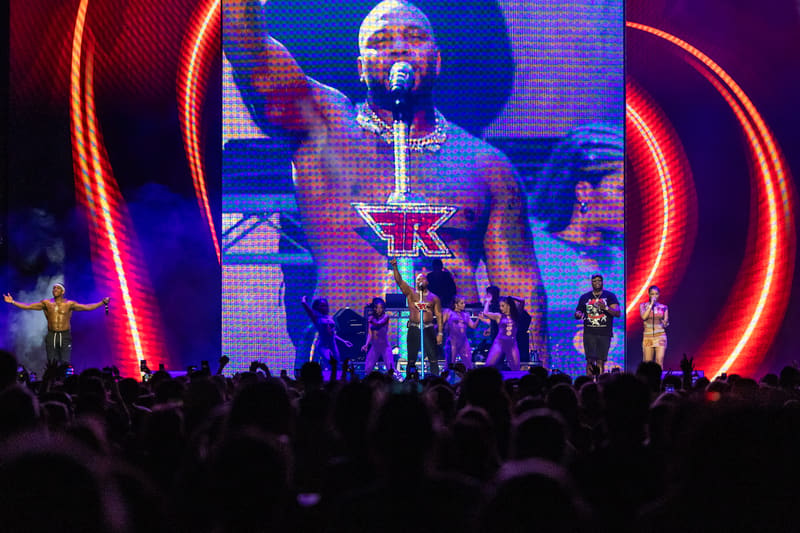 Performer on stage beneath large video screen with red neon curved light installations, surrounded by crowd silhouettes.