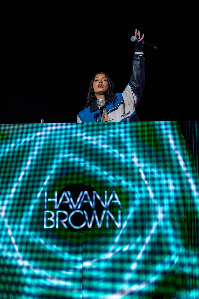 Performer at a DJ booth raising one arm to the crowd above a glowing cyan stage screen displaying "HAVANA BROWN".