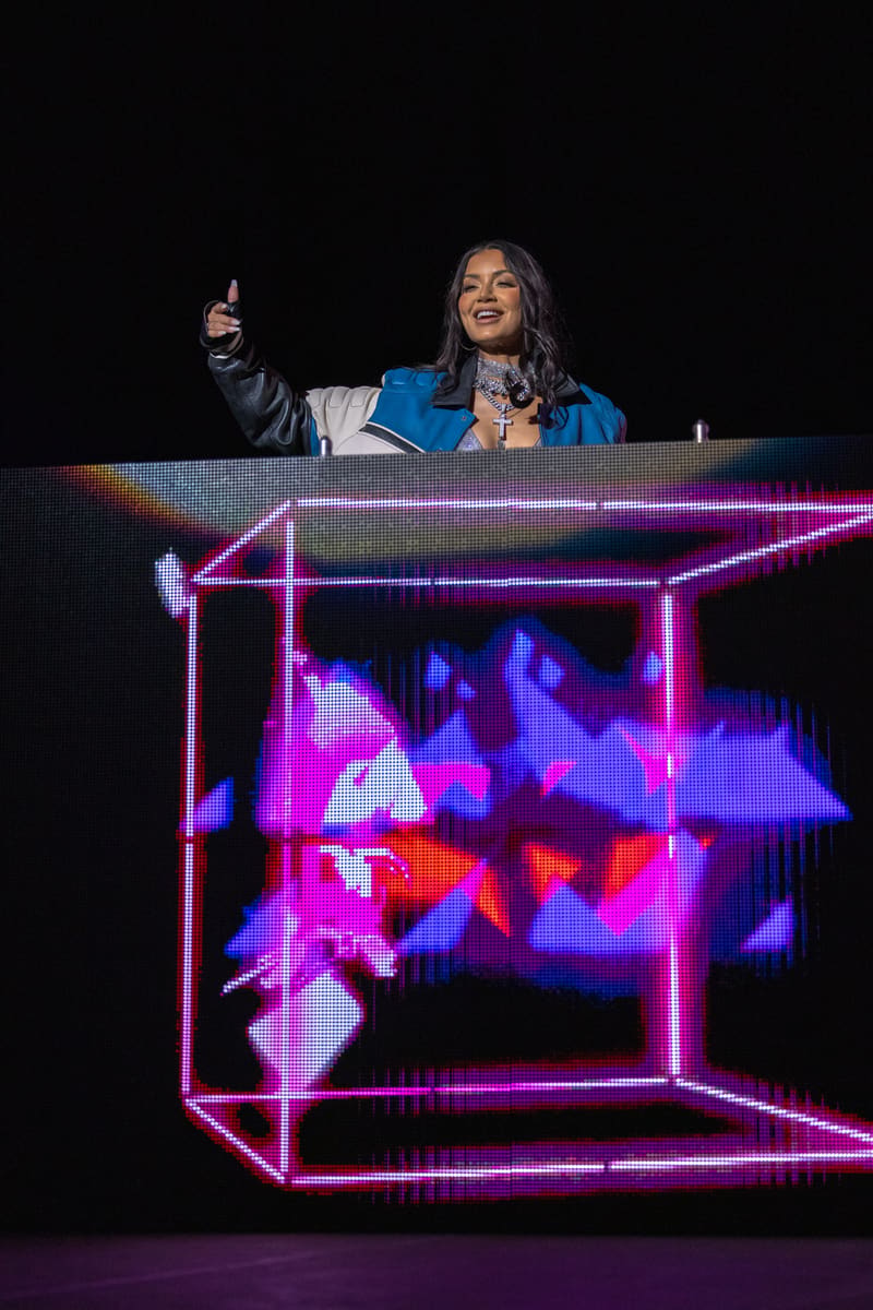 Performer at DJ booth with neon pink geometric frame and video screens displaying dancers behind her on stage.