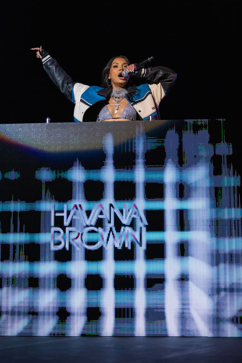 Havana Brown performs at a DJ booth with raised arm, illuminated by blue and white LED screen displaying her name.
