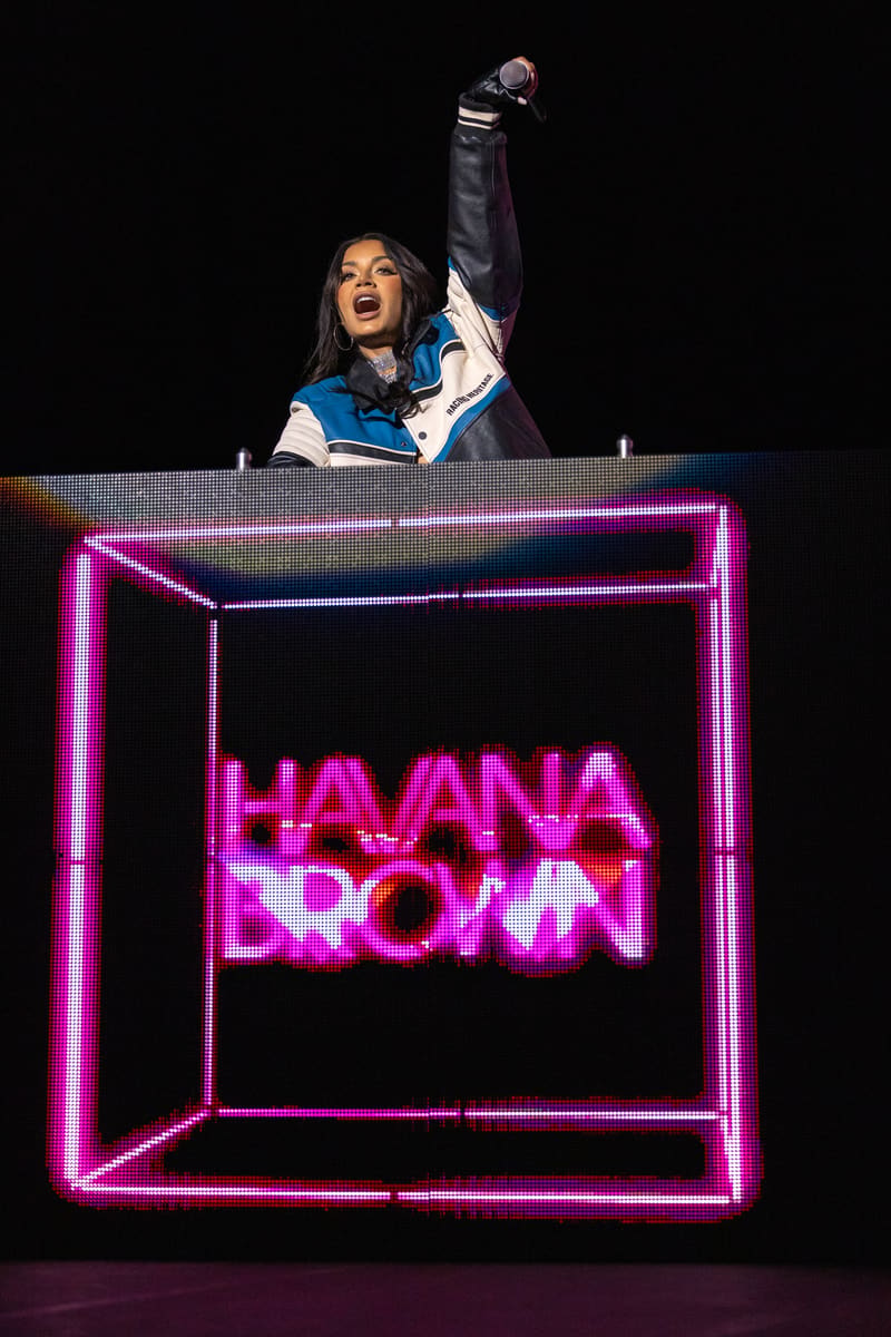 Performer at DJ booth with fist raised, wearing blue and white jacket, above neon-pink "HAVANA BROWN" sign.
