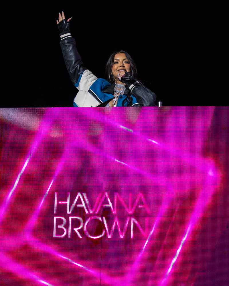 Performer on stage with raised fist, backed by glowing magenta neon signage reading "HAVANA BROWN".