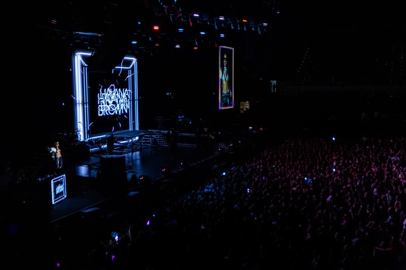 Performer on a dimly lit stage with blue neon signage, facing a large crowd in a dark venue.