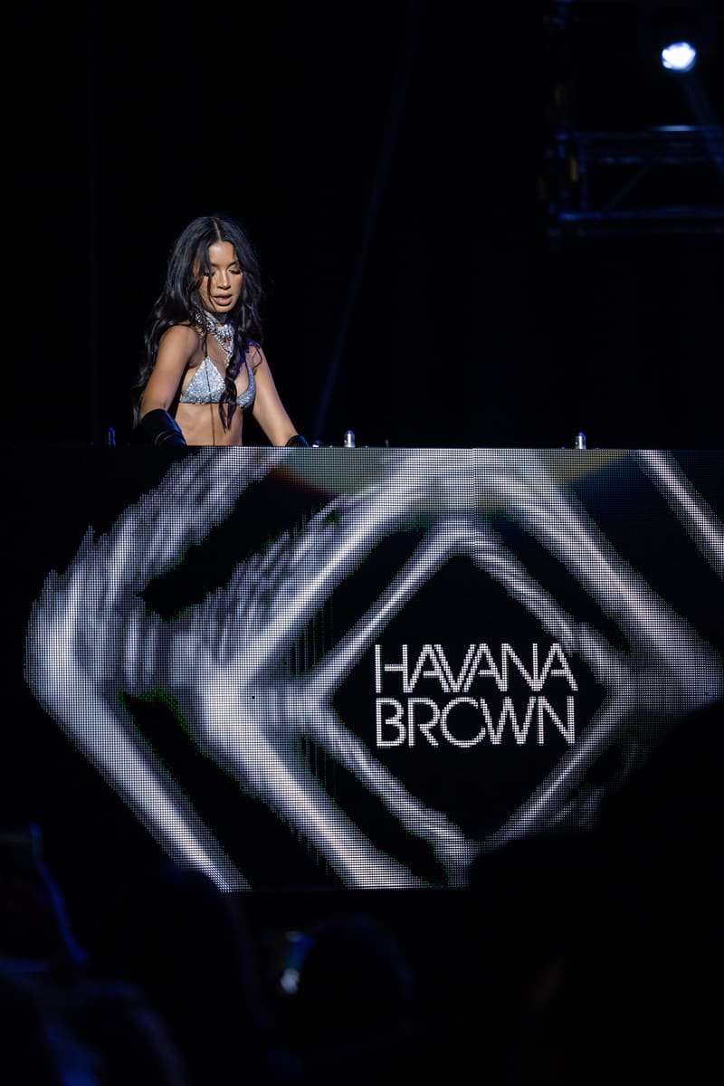 DJ Havana Brown at turntables beneath animated visual projection on dark stage.
