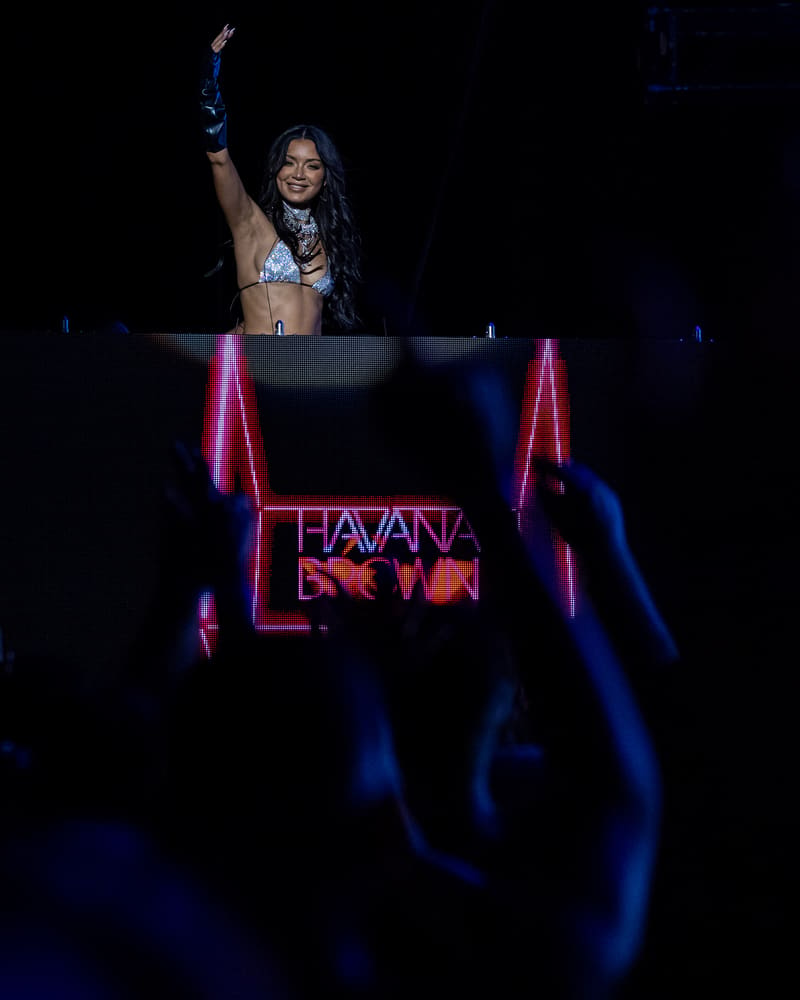 DJ Havana Brown raises her fist at the decks, surrounded by neon pink signage and blue stage lighting.