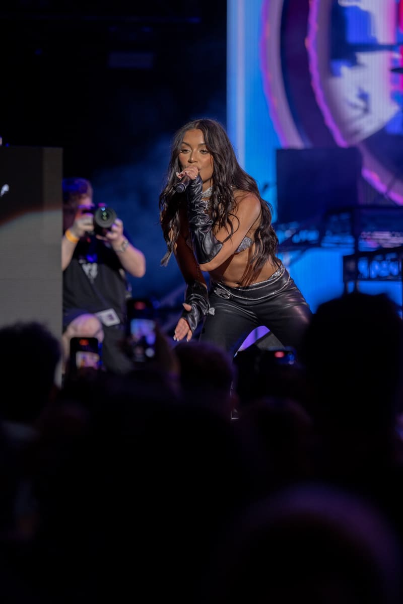 Performer singing into microphone on stage under blue and purple lights, audience members recording with phones in foreground.